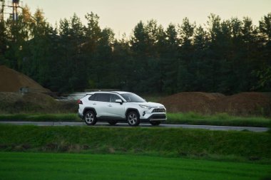 Melez bir SUV arabası görkemli bir doğa ve arka planda altın bir günbatımına sahip manzaralı bir yolda ilerliyor..