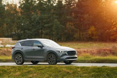 Melez bir SUV arabası görkemli bir doğa ve arka planda altın bir günbatımına sahip manzaralı bir yolda ilerliyor..