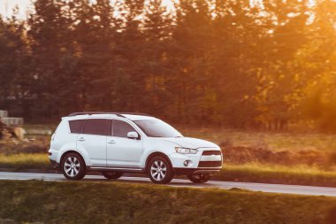 Melez bir SUV arabası görkemli bir doğa ve arka planda altın bir günbatımına sahip manzaralı bir yolda ilerliyor..