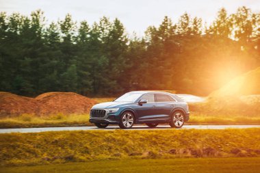 Melez bir SUV arabası görkemli bir doğa ve arka planda altın bir günbatımına sahip manzaralı bir yolda ilerliyor..