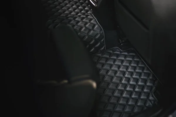 A sophisticated and luxurious leather floor mat in the second row of a modern car. The interior is black and elegant, with a dashboard that has technology and safety features, a seat, and pedals.