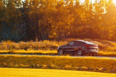 Melez bir SUV arabası görkemli bir doğa ve arka planda altın bir günbatımına sahip manzaralı bir yolda ilerliyor..