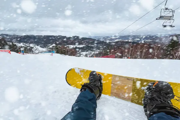Snowboarding POV. First-person view of the snowy slope and the mountains. The sky is cloudy and white, creating a serene atmosphere.