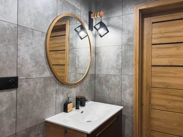 A Sleek and Simple Bathroom with a Wooden Door and an Oval Mirror. The bathroom has a grey tiled wall and a white sink with a faucet. The soap dispenser is also white and contrasts with the wood.