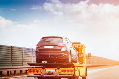 Siyah bir SUV, üzerinde bulutlu bir gökyüzü olan boş bir otoyol boyunca sarı bir çekiciyle taşınıyor..