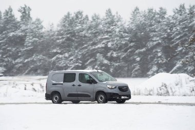Kışın karlı bir yolda kargo taşıyan bir minibüs. Kar ve buzla kaplı bir yolda giden mavi bir minibüsün yan görüntüsü. Yol karlı bir manzara ve bulutlu bir gökyüzü ile çevrilidir..