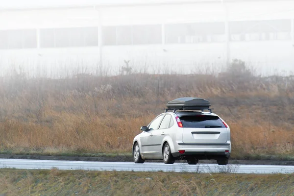 A modern car with a roof box travels down a road surrounded by grasslands under an overcast sky.