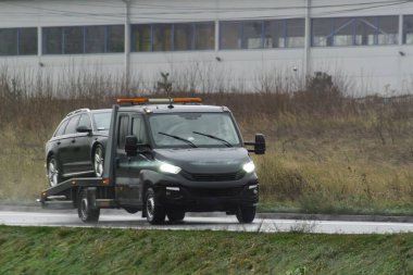 Bir kamyon arızalı bir araba taşıyor. Arabanın motorunda bir sorun var ve olay yerinde tamir edilemez. Çekici servisi arabayı tamirciye teslim ediyor.