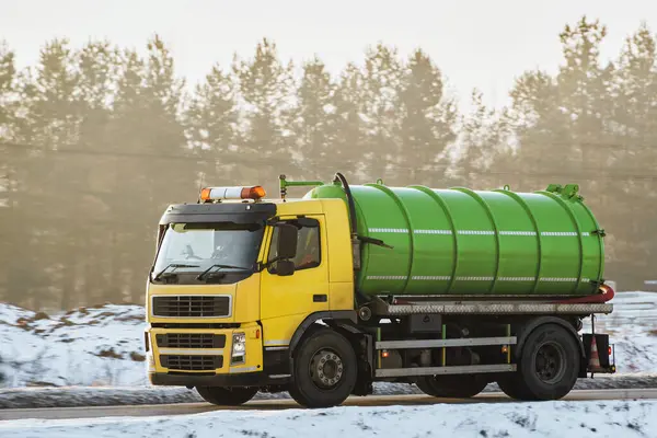 A yellow septic truck with a green tanker is moving on a snowy road amidst trees as the sun sets casting a warm glow. Vacuum truck in Europe.