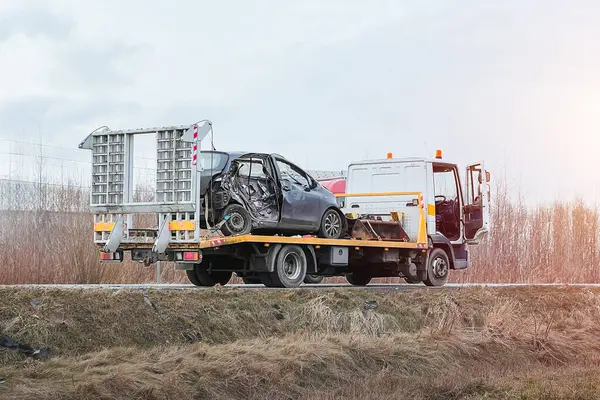 A side-impact collision leaves a car in need of a tow truck.  The car is severely damaged and may be beyond repair. A modern black Japanese hatchback is being hauled away by a tow truck on the highway
