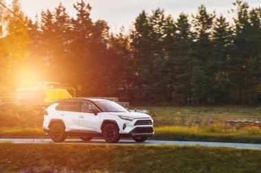 Melez bir SUV arabası görkemli bir doğa ve arka planda altın bir günbatımına sahip manzaralı bir yolda ilerliyor..