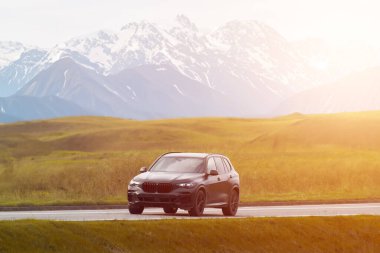 Siyah lüks SUV dağlarla dolu manzaralı kırsal alanda ilerliyor.