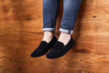 Siyah süet ayakkabılar ve ahşap arka planda oturan mavi kot giyen bir adamın yakın çekimi. Görüntü tarzı ve konforu çağrıştırıyor.