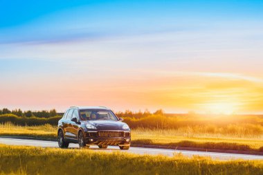 Şatafatlı siyah SUV gün batımında bir kır yolunda sürüyor.