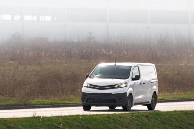 Arka planda sonbahar yeşilliği olan güneşli bir otoyolda hızla giden modern bir teslimat aracı.