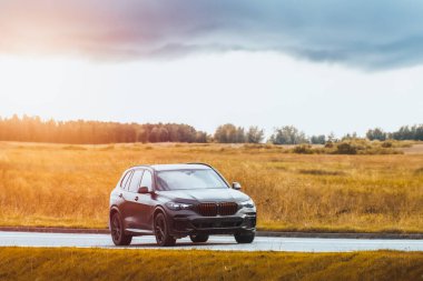 Pahalı, siyah arazi aracı yeşil tarlalardan geçen manzaralı bir yolda gidiyor.