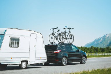 Car with bicycles mounted on top towing a caravan on a sunny highway against a scenic mountain backdrop, family road trip adventure during vacation clipart