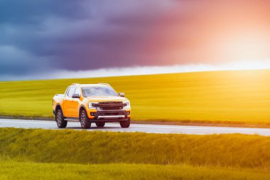 Turuncu kamyonet kırsal bir yolda dramatik bir günbatımı gökyüzüne doğru ilerliyor.