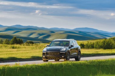 Alman, lüks siyah SUV gün batımında bir kır yolunda ilerliyor, arka planda tepeler yuvarlanıyor.