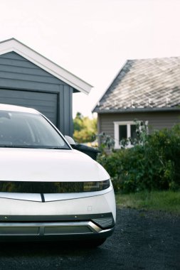 Gri bir garajın önüne park edilmiş beyaz elektrikli araba.