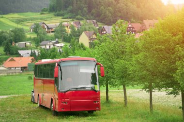 Arka planda köy evleri olan ağaçların arasına park edilmiş kırmızı otobüs.