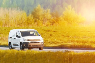 Beyaz minibüs manzaralı sonbahar kırsalından geçiyor.
