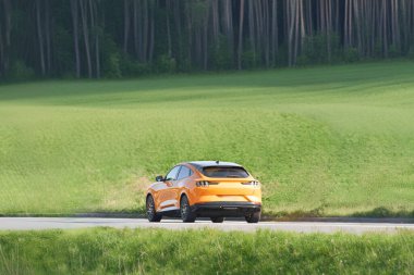 Modern electric orange SUV drives through a peaceful countryside, surrounded by lush forest