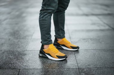 Modayı takip eden sarı ve siyah spor ayakkabılı, ıslak şehir kaldırımında duran koyu renk kot pantolonlu, kentli sokak tarzını ve aktif yaşam tarzını gösteren kendine güvenen bir adam.