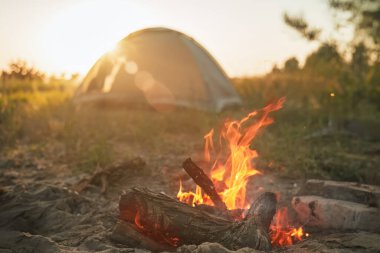 Gün batımında arka planda bir çadırla kamp ateşi yanıyor.
