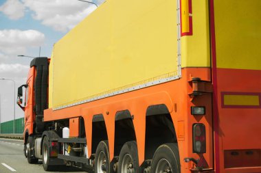 Large orange semi truck is driving down a highway on a sunny day. The back of the truck has space for branding or advertising clipart