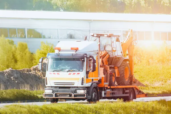 Turuncu çekici yazın kırsal bir yolda mini kazıcı taşıyor.