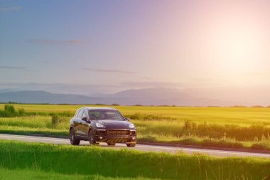 Alman siyah SUV gün batımında tarlaları ve tepeleriyle bir kır yolunda ilerliyor.