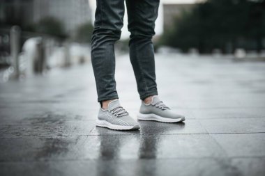 Havalı gri spor ayakkabılarıyla ıslak şehir kaldırımında dikilen şık bir adam gündelik ve modern ayakkabılarına dikkat çekiyor. Yakın plan çekimler rahat ayakkabı ve kot pantolonlar, şehir tarzı için mükemmel.