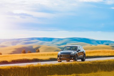 Alman, lüks siyah SUV gün batımında bir kır yolunda ilerliyor, arka planda tepeler yuvarlanıyor.