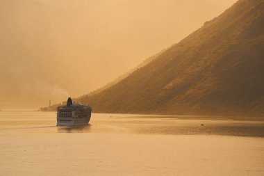 Gün batımında görkemli dağların yakınlarındaki sakin sularda seyreden bir gemi.