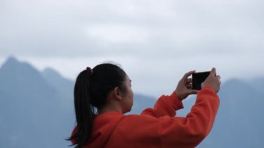 Sakin bir dağın tepesinde duran genç bir kadının arka görüntüsü ve telefonda güneşin doğuşundaki güzel dağ manzarasının fotoğraflarını çekiyor..