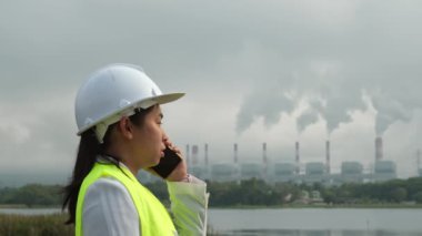 Yeşil yelek ve miğfer giyen kadın mühendis dışarıda, kömür santralinin arka planında ve sabah sisinde buharla konuşuyor..