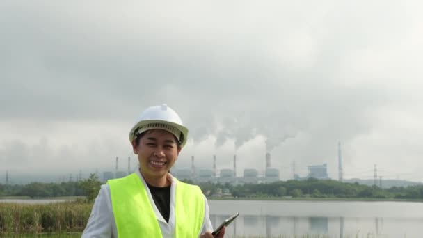 朝の霧の中で石炭や蒸気発電所の背景を背景に外に立って緑のベストとヘルメットで笑顔の女性チーフエンジニア 石炭火力発電所で働くエンジニアとラップトップ — ストック動画