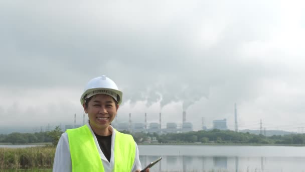 緑のベストとヘルメットを着た女性チーフエンジニアは 朝の霧の中で石炭発電所や蒸気の背景に外に立っています 石炭火力発電所で働くエンジニアとラップトップ — ストック動画