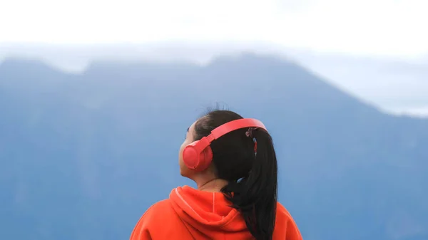 穏やかな丘の上に立って 午前中にヘッドフォンで音楽を聴く若い女性の後部ビュー 冬には山を眺めながら自然の美しさを楽しむセーターを着た女性 — ストック写真