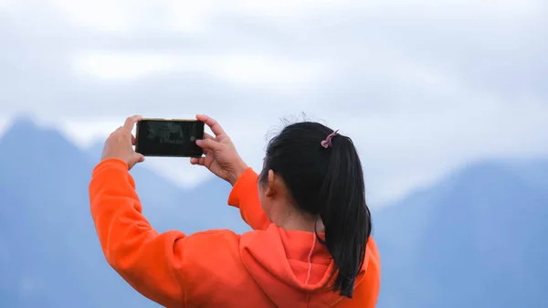 Rear view of a young woman standing on the top of a calm mountain and taking pictures of the beautiful mountain scenery at sunrise on the phone.