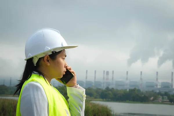 stock image Female chief engineer in green vest and helmet talking on the phone outside against the background of coal power plant station and steam in the morning mist.