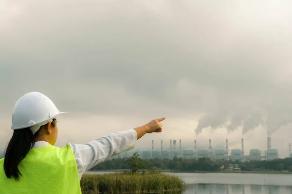 stock image Female chief engineer wearing a green vest and helmet stands outside against the background of coal power plant station and steam in the morning mist. Engineer working and laptop at coal power plant.