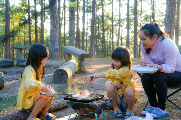Happy mother and children picnic sit by stove near tent and grill a barbecue in pine forest, eating and have conversation. Happy family on vacation in nature.