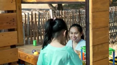 Happy sisters play with food and wooden grocery toys at the outdoor playground with her mother. Cute Asian girl roleplaying selling fruit juice at the park. Family spending time together on vacation.