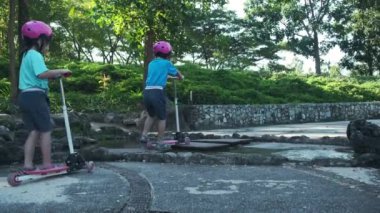 Portrait of active little girl riding scooter on street in outdoor park on summer day. Happy Asian girl wearing a helmet riding a kick scooter in the park. Active leisure and outdoor sport for child.