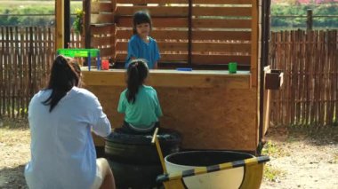 Happy sisters play with food and wooden grocery toys at the outdoor playground with her mother. Cute Asian girl roleplaying selling fruit juice at the park. Family spending time together on vacation.