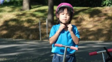 Portrait of young active girl wearing helmet getting ready to ride scooter on road in outdoor park on summer day. Happy Asian girl riding a scooter to school.