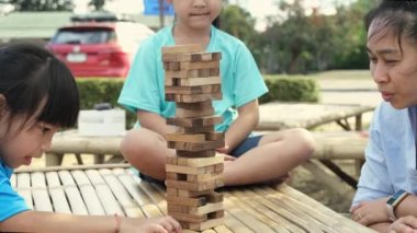 Excited kids and mom playing Jenga tower wooden block game together in the park. Happy family with children enjoying weekend activities together.