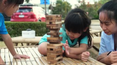 Excited kids and mom playing Jenga tower wooden block game together in the park. Happy family with children enjoying weekend activities together.